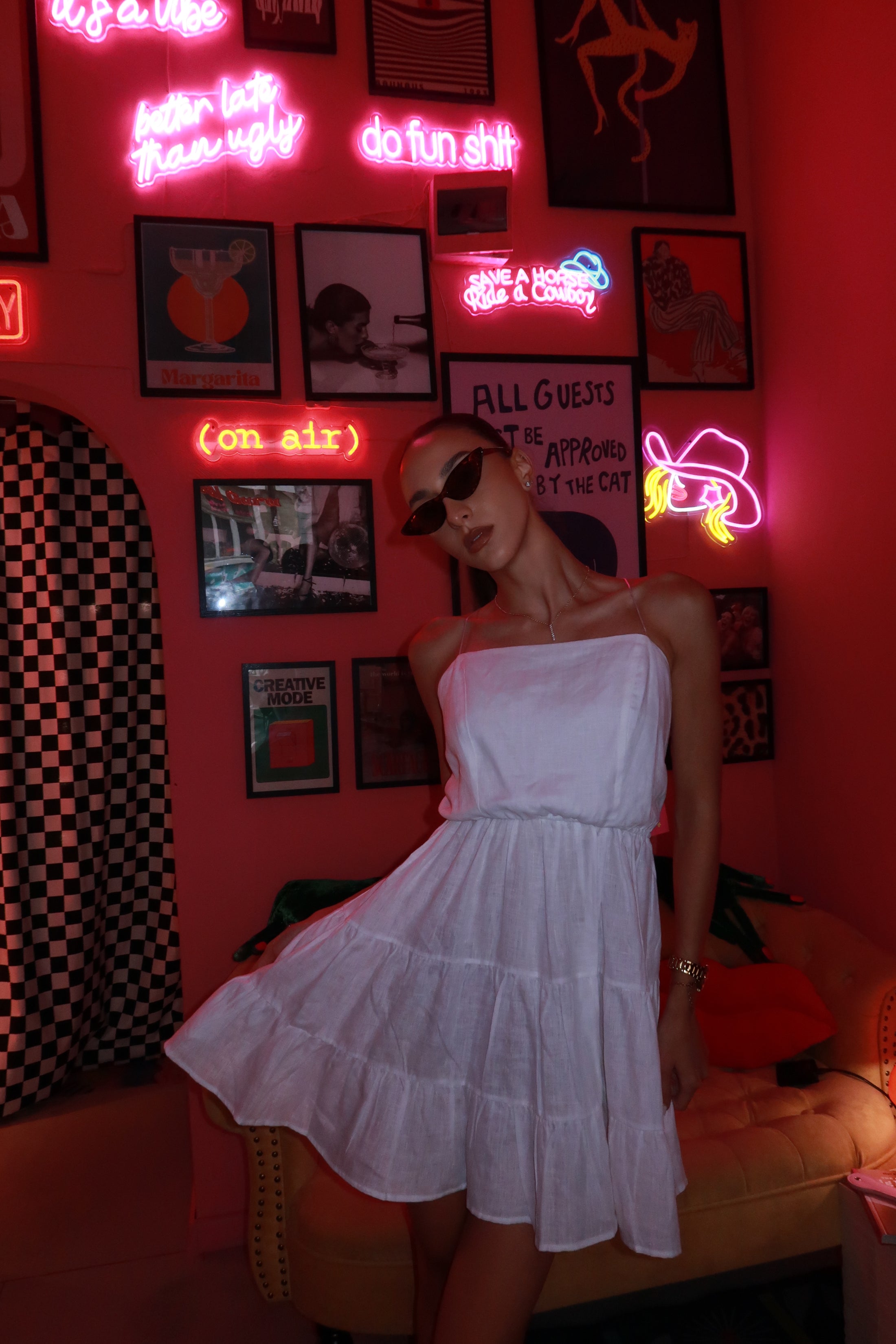Woman in a white dress standing in a room with neon signs and posters on a red wall.