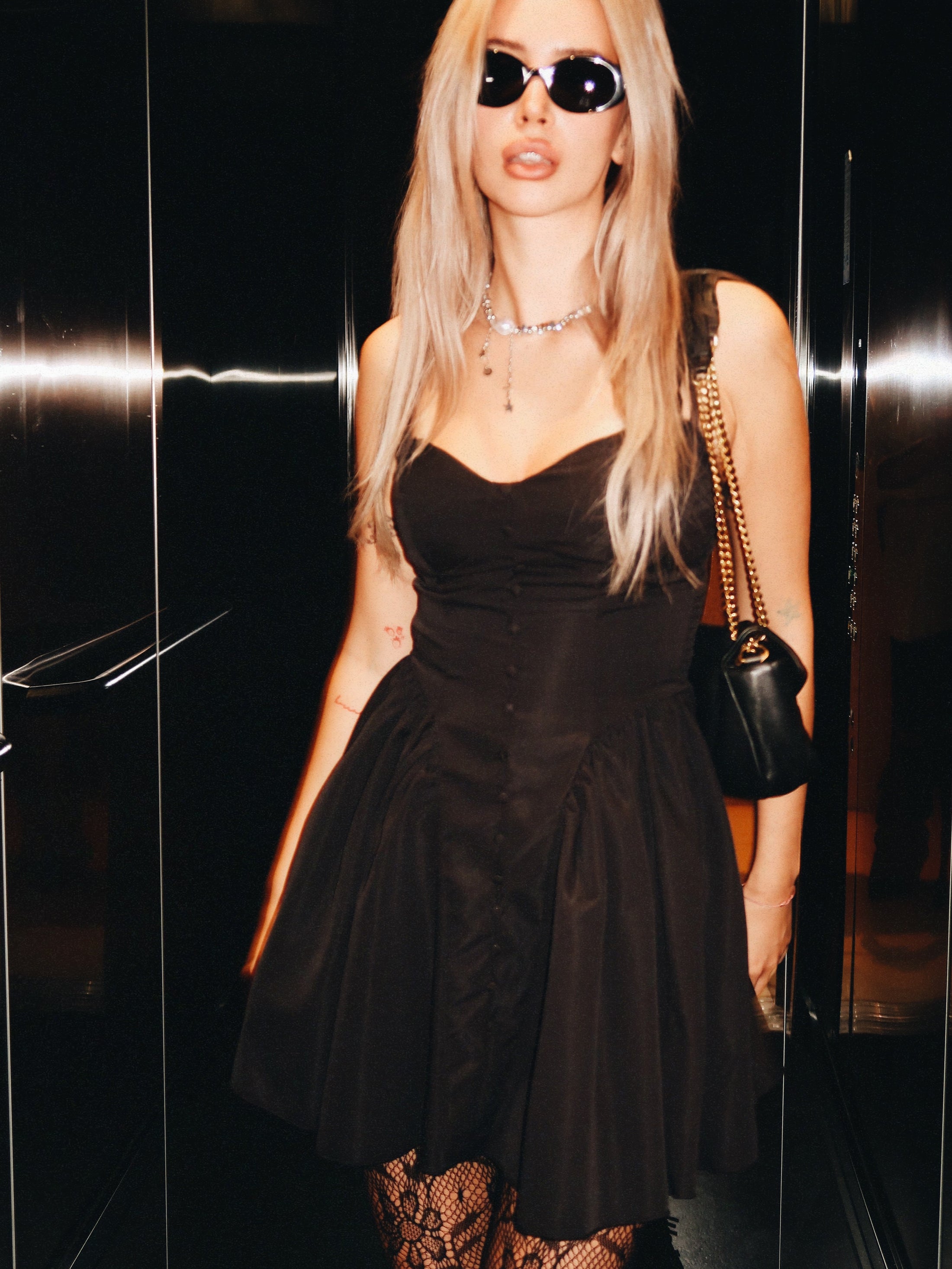 Woman wearing a black dress and sunglasses in front of a reflective surface