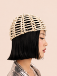 Load image into Gallery viewer, A woman in profile wearing a beige crochet or knit beaded hat with a black bob haircut, set against a solid pale peach background.