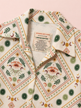 Load image into Gallery viewer, A close-up of a collared button-up shirt with a cream base and a detailed bohemian-style print featuring green, orange, and brown celestial motifs like crescent moons, stars, and eye designs. The inner collar label reads 'STYLANE ART X KIKI LIMITED EDITION'.