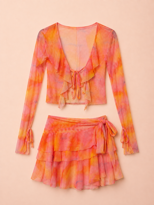 A two-piece women's outfit set featuring a long-sleeve ruffled crop top and a matching tiered ruffled mini skirt, both in a vibrant orange and pink tie-dye pattern, displayed against a solid pale peach background.