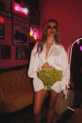 Load image into Gallery viewer, A person wearing a white satin crop shirt with long sleeves, styled with a knot and twist detail. The shirt is paired with a green mini skirt and accessorized with sunglasses and a watch.