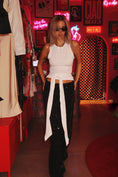 Заредете изображението в програмата за преглед на галерия, Woman in a white top and black pants with a belt standing in a neon-lit room.