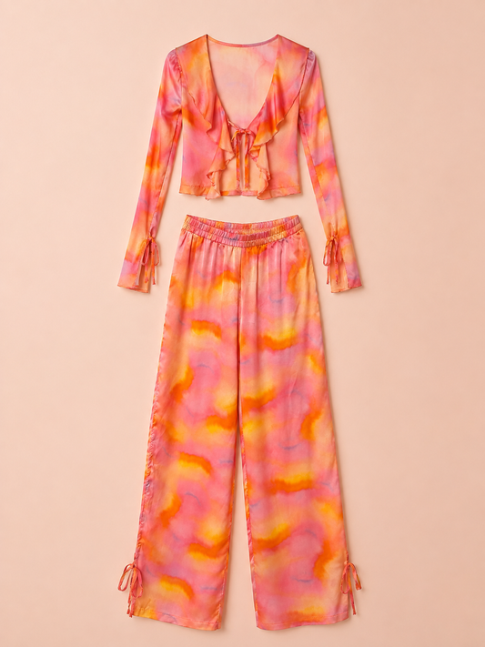 A two-piece matching set featuring a long-sleeve crop top with a ruffled V-neckline and tie-dye pattern, paired with wide-leg pants with side tie details, shown flat on a solid peach background.