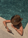 Load image into Gallery viewer, A person sitting by a swimming pool wearing a brown woven cowboy hat with a silver skull and band detail, along with a dark mesh top and bracelets.