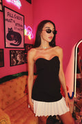 A person wearing a strapless black dress with white pleated mini skirt, standing in a room with pink walls and decorative lights.