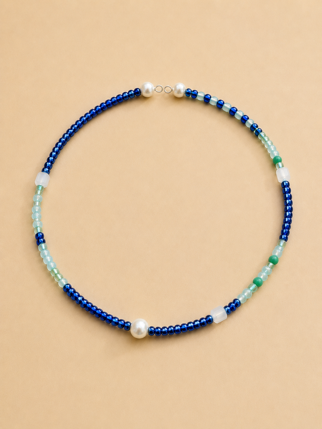 A beaded necklace with alternating strands of deep blue beads, light aqua beads, white translucent beads, and a single white pearl, displayed on a solid beige background.