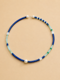 A beaded necklace with alternating strands of deep blue beads, light aqua beads, white translucent beads, and a single white pearl, displayed on a solid beige background.