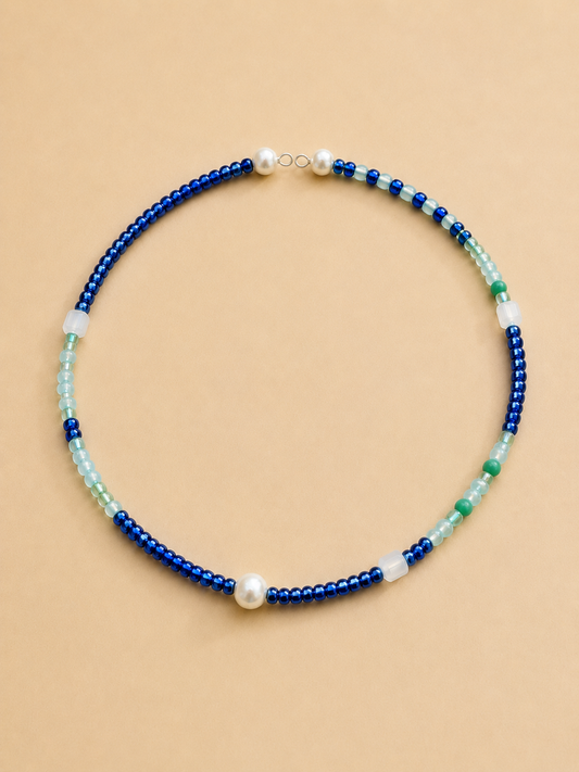 A beaded necklace with alternating strands of deep blue beads, light aqua beads, white translucent beads, and a single white pearl, displayed on a solid beige background.