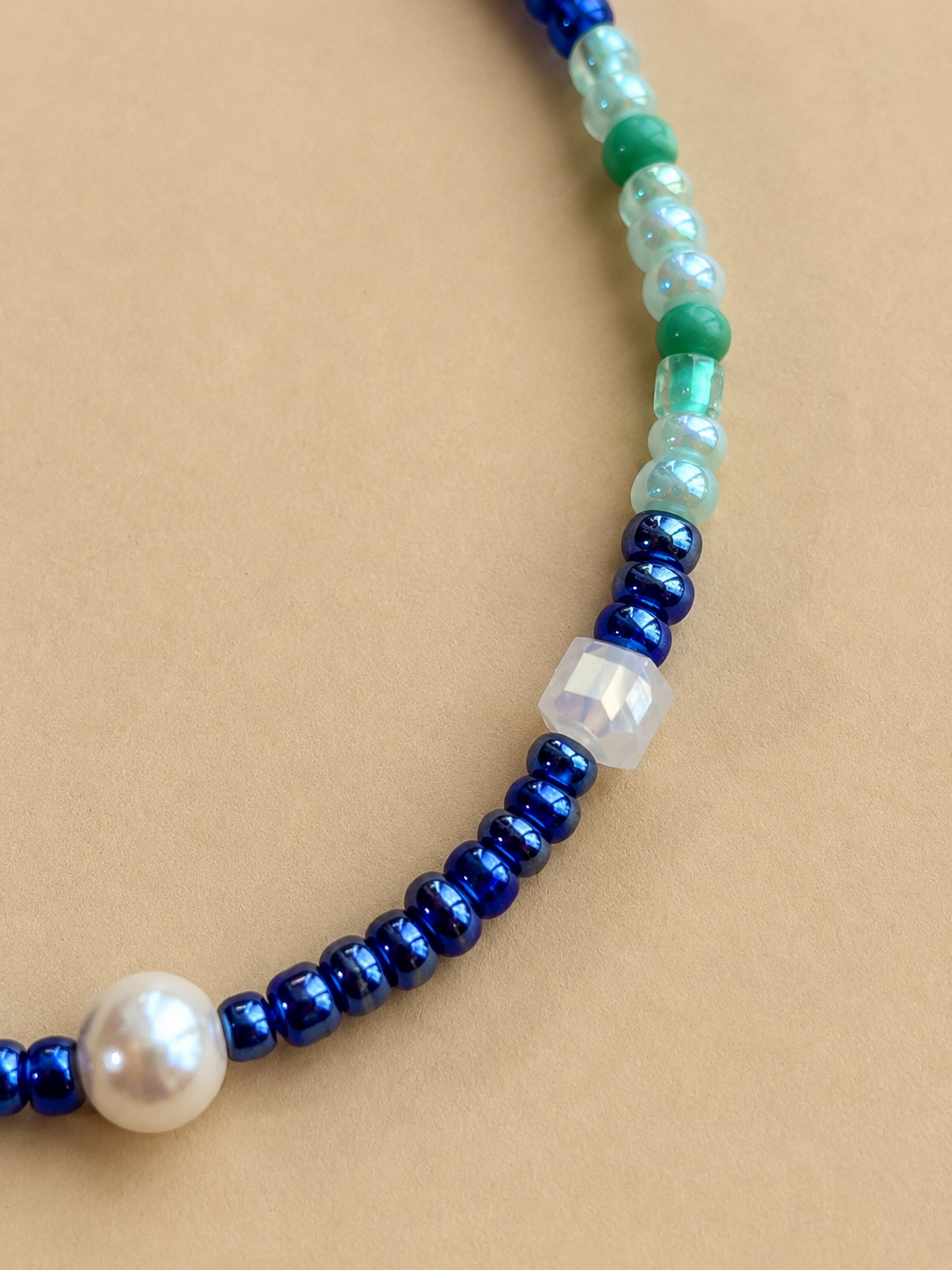 A close-up of a beaded necklace featuring metallic royal blue beads, a white pearl, and translucent green and iridescent beads, laid on a solid tan background.
