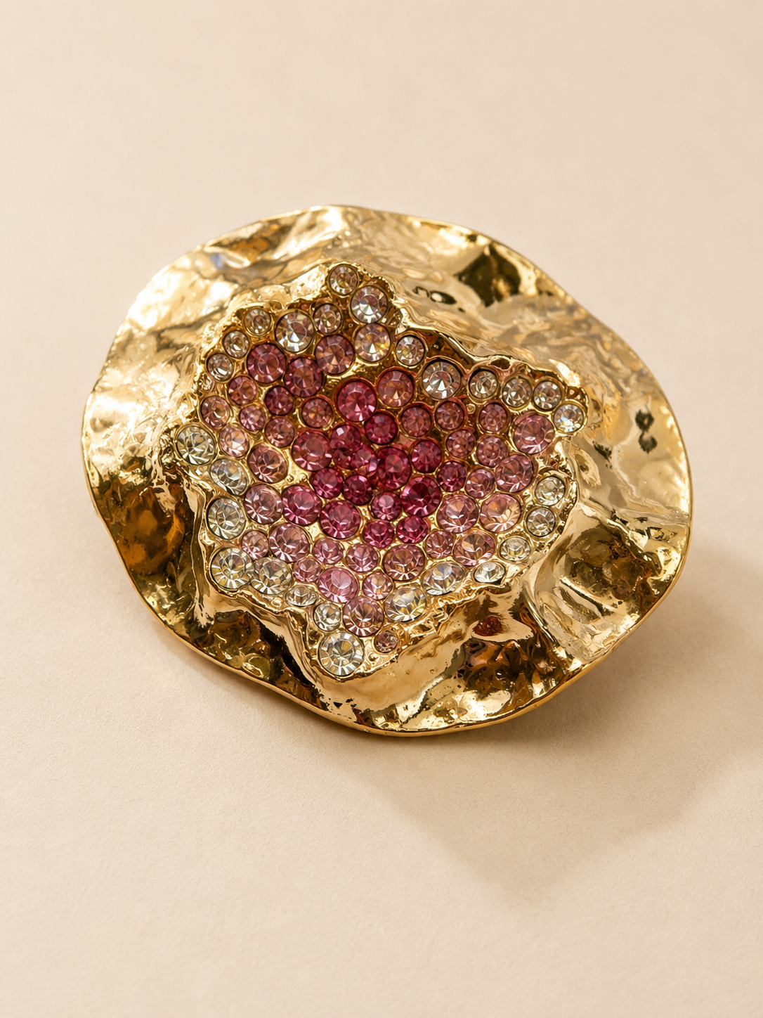 A gold-toned, organic-shaped earring featuring a central cluster of pink and clear gemstones, photographed on a solid beige background.