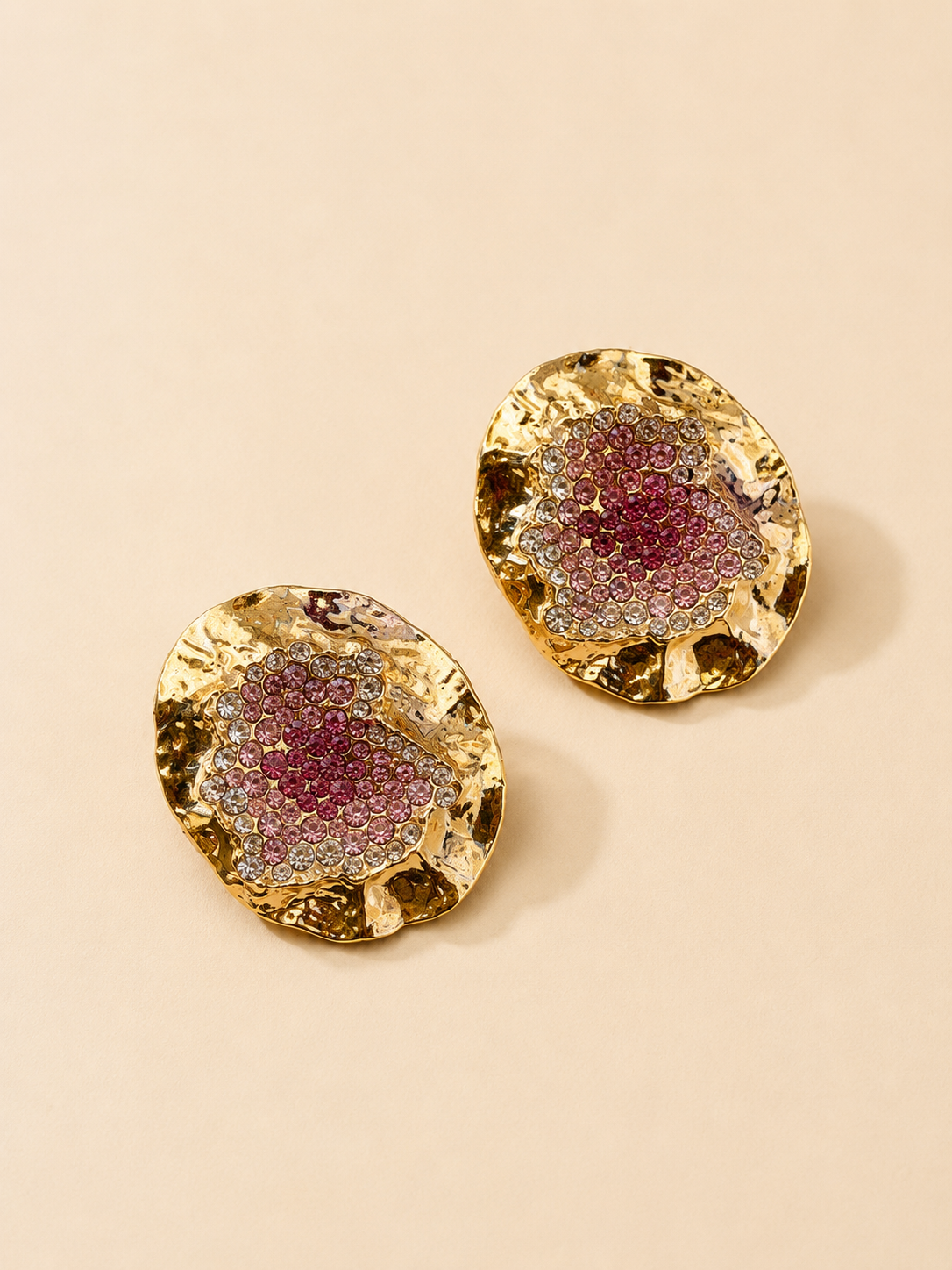A pair of gold-toned disc-shaped earrings with textured, organic edges and a central cluster of sparkling pink and silver rhinestones, displayed on a warm beige background.