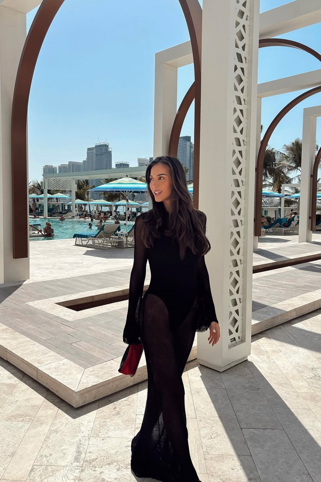 Woman in a black outfit standing in front of a pool area with buildings and palm trees in the background.
