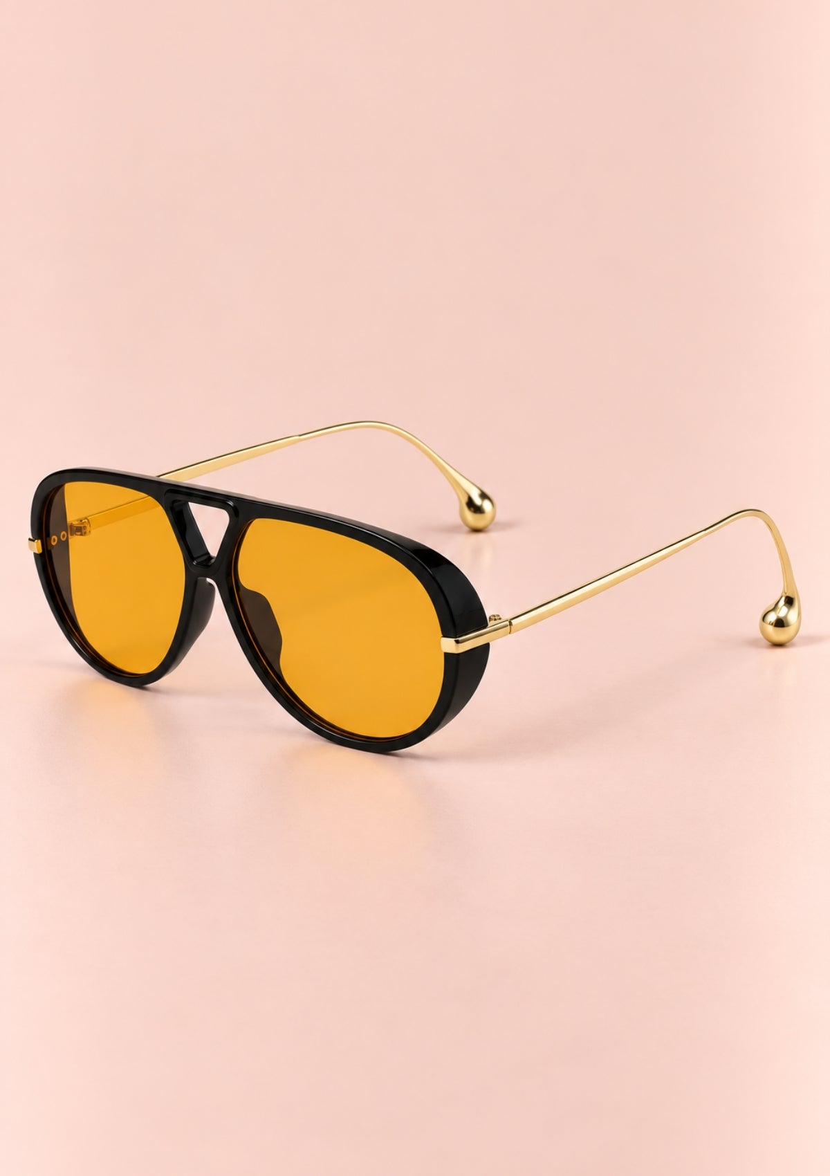 Aviator-style sunglasses with a glossy black frame and bright yellow lenses, featuring slim gold metal temples with decorative bead tips, set against a solid pale pink background.