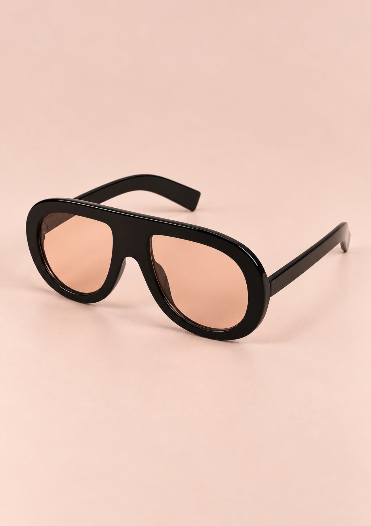 Black aviator-style sunglasses with oversized black frames and warm pink-tinted lenses, shown on a solid pale pink background.