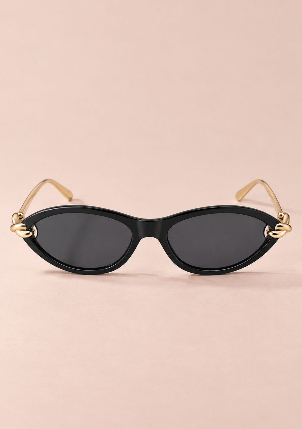 Oval sunglasses with a black frame, dark lenses, and gold metal temples with decorative knot details at the hinges, shown on a solid pale pink background.