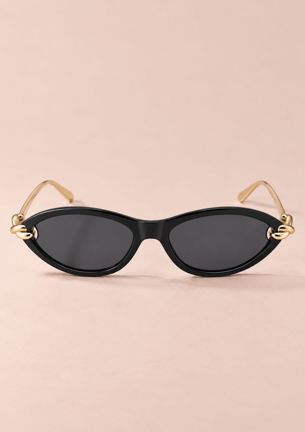 Oval sunglasses with a black frame, dark lenses, and gold metal temples with decorative knot details at the hinges, shown on a solid pale pink background.