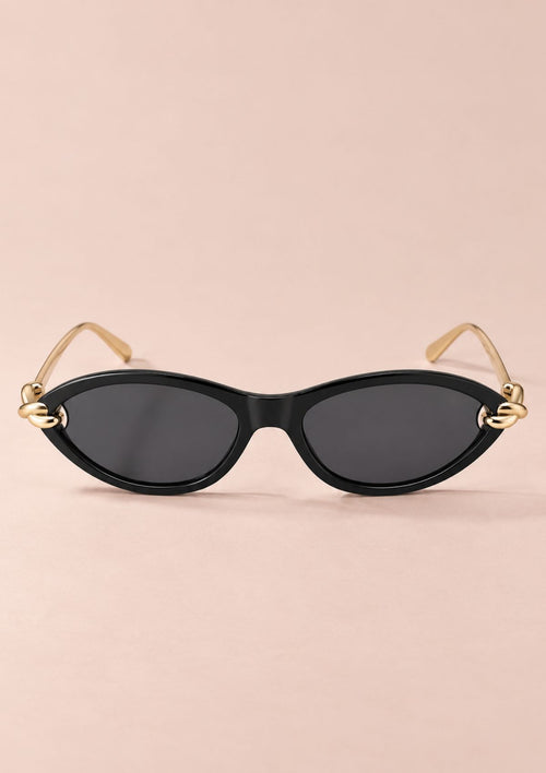 Oval sunglasses with a black frame, dark lenses, and gold metal temples with decorative knot details at the hinges, shown on a solid pale pink background.