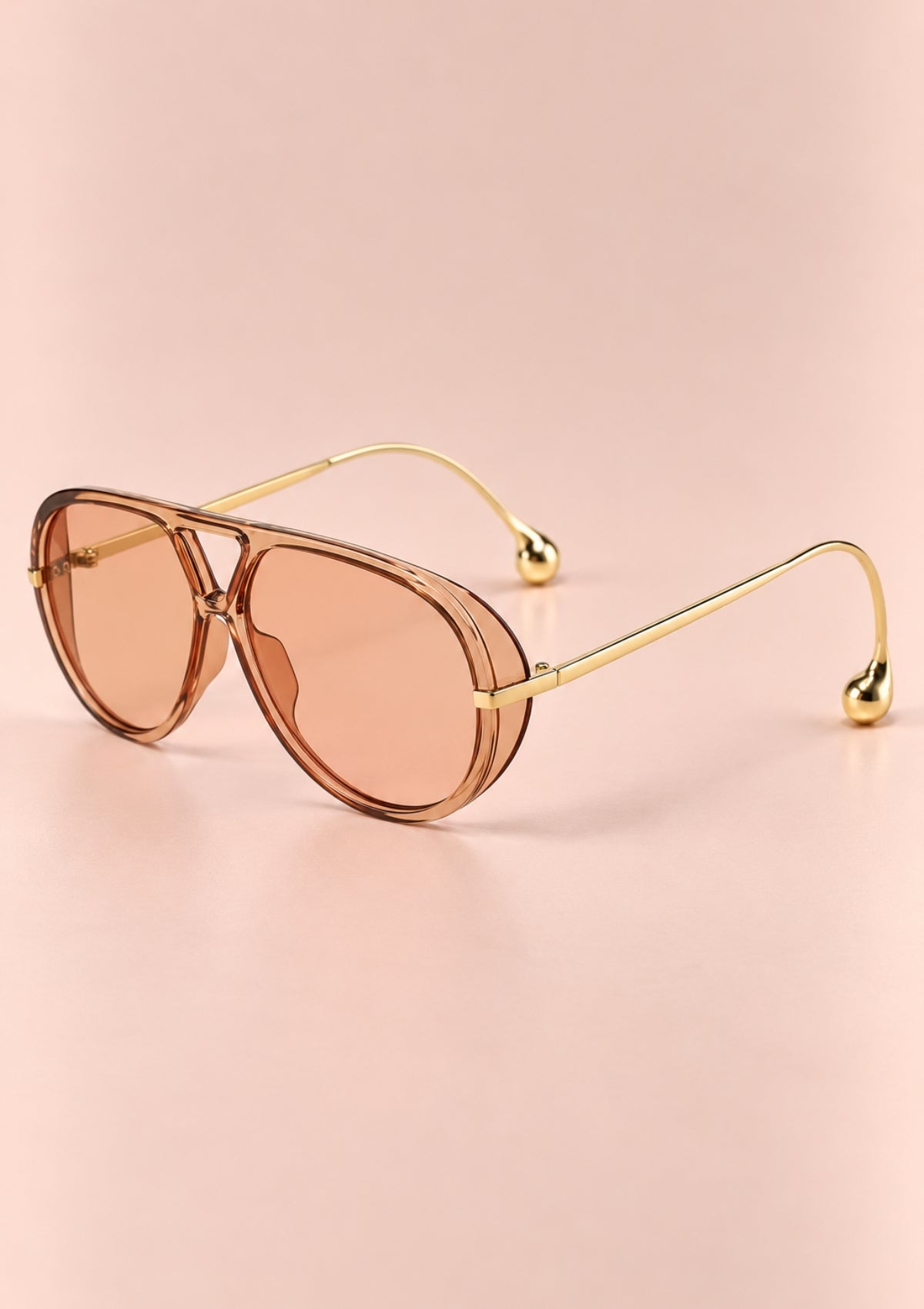Aviator-style sunglasses with a gold metal frame and translucent pink-tinted lenses, displayed on a soft pink background.
