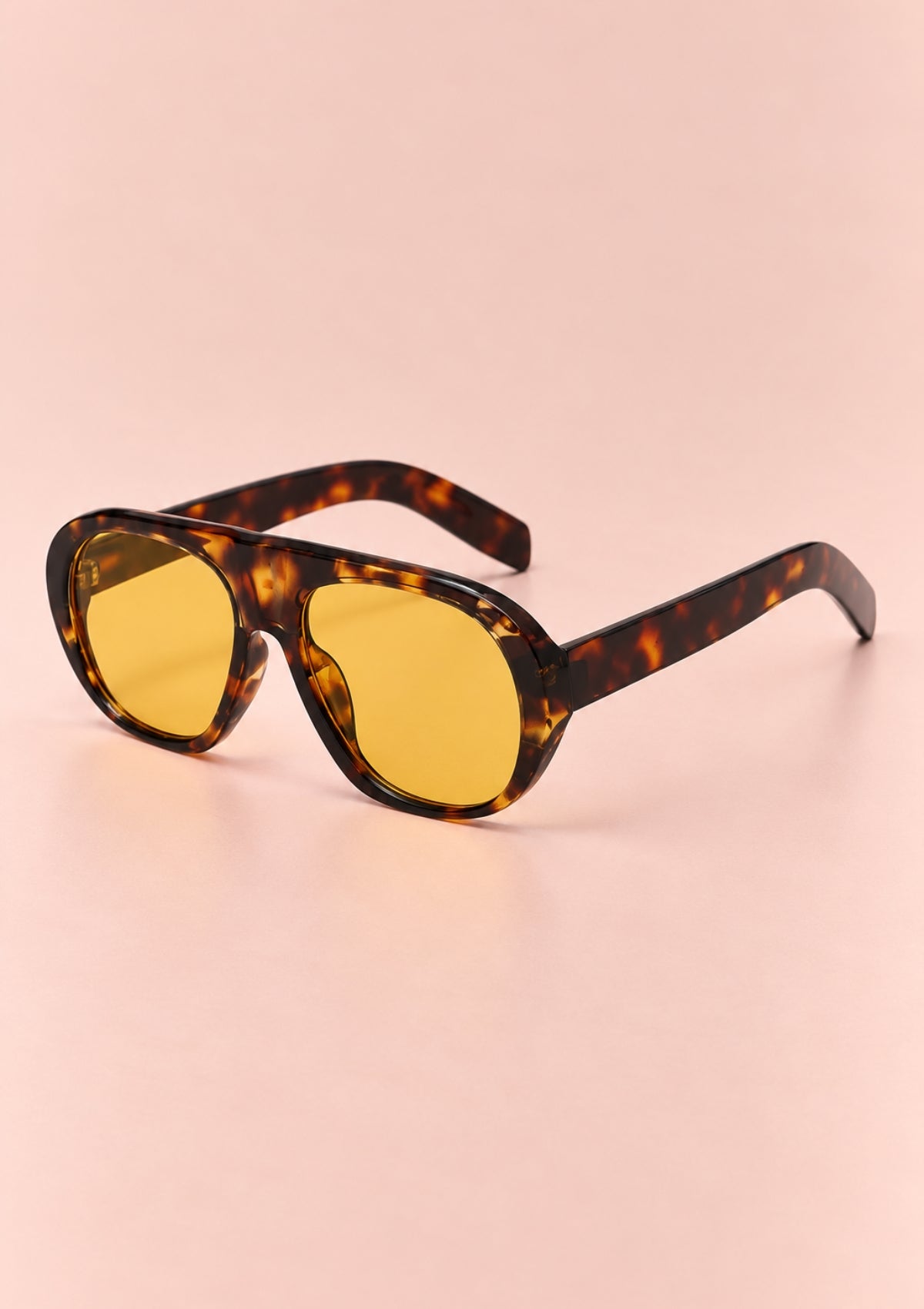Oversized shield-style sunglasses with a tortoiseshell frame and yellow tinted lenses, photographed on a soft peach-pink background.