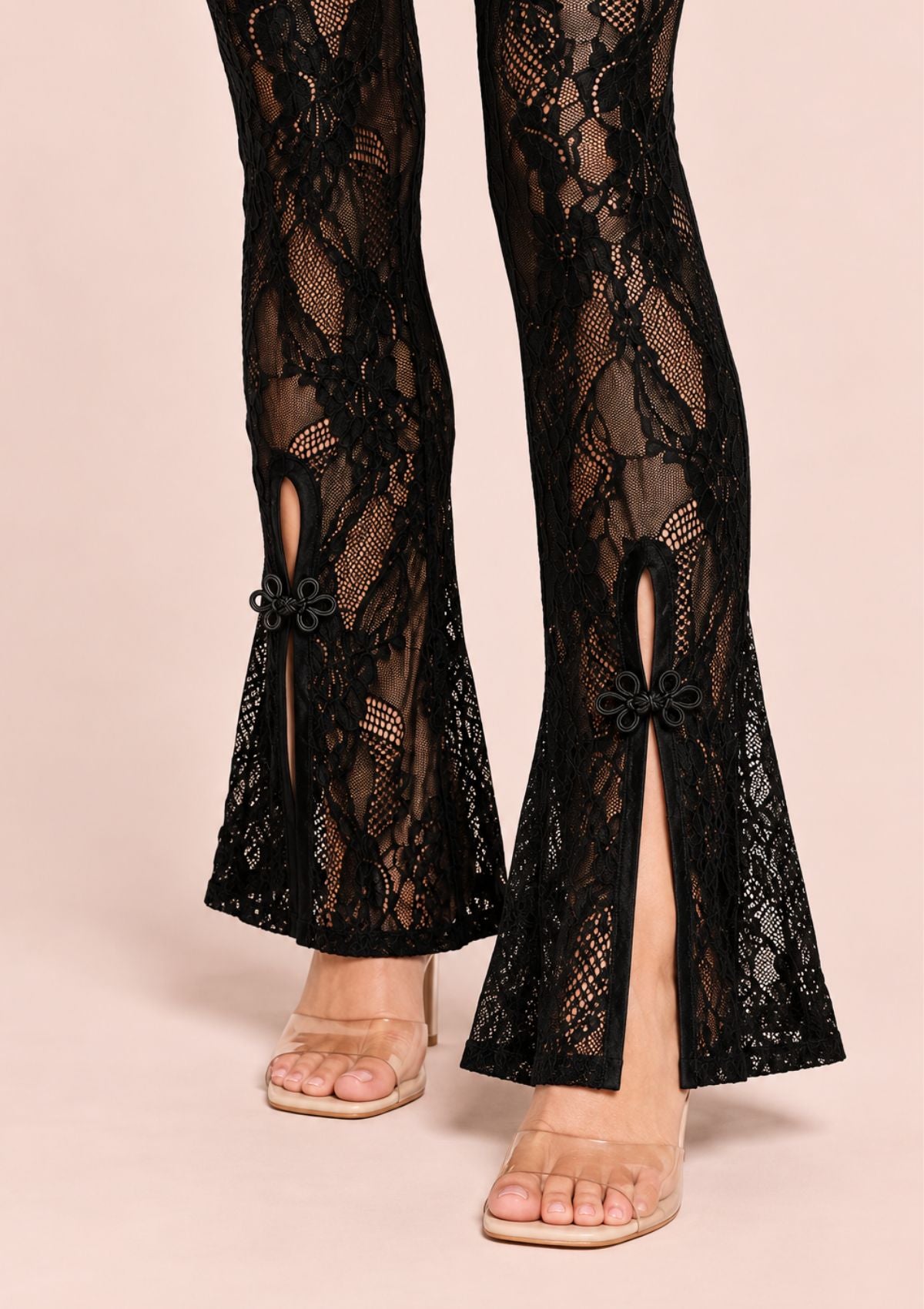 Black lace flare pants with side slit details and small bow accents, shown on a model wearing clear-strap heels against a solid pale pink background.