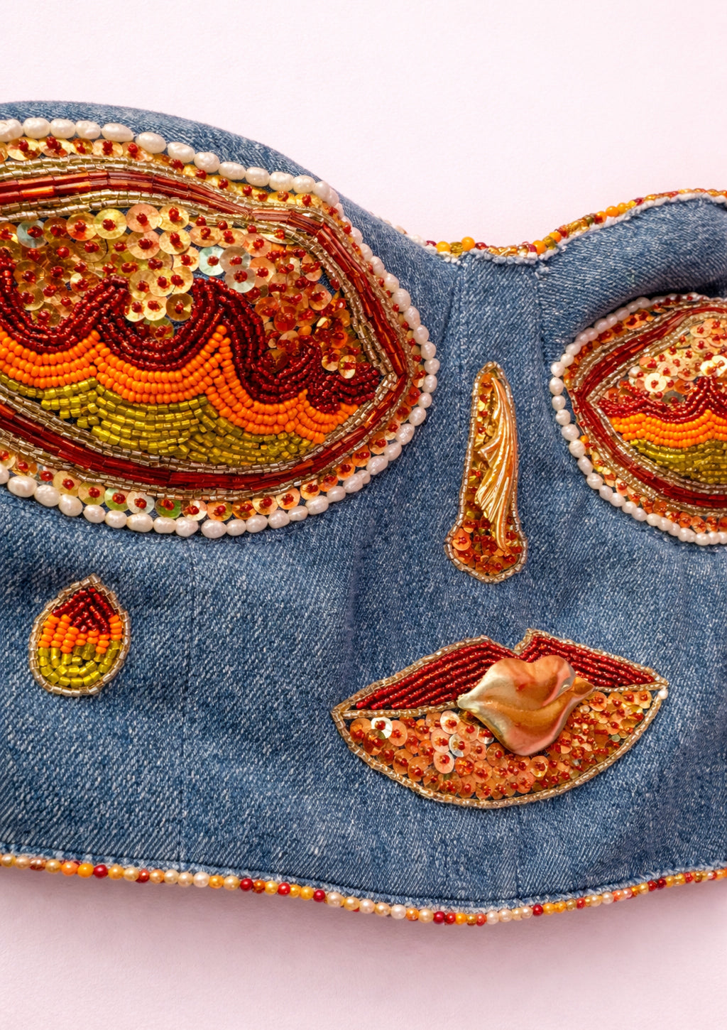 Decorative denim eye corset with bead and fabric embellishments on a light pink background