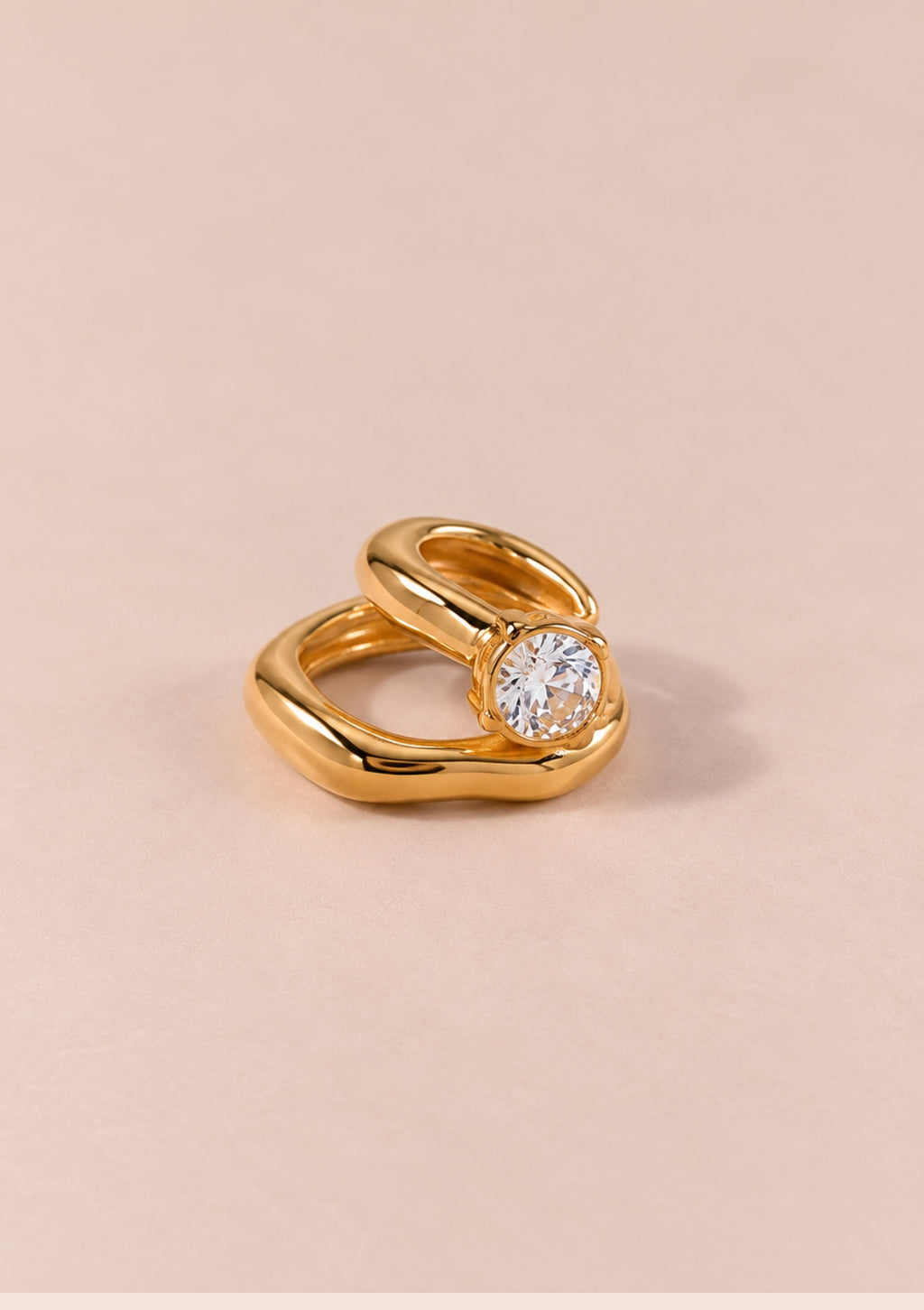 A gold ring with a twisted, fluid design and a round clear gemstone, photographed on a solid pale pink background.
