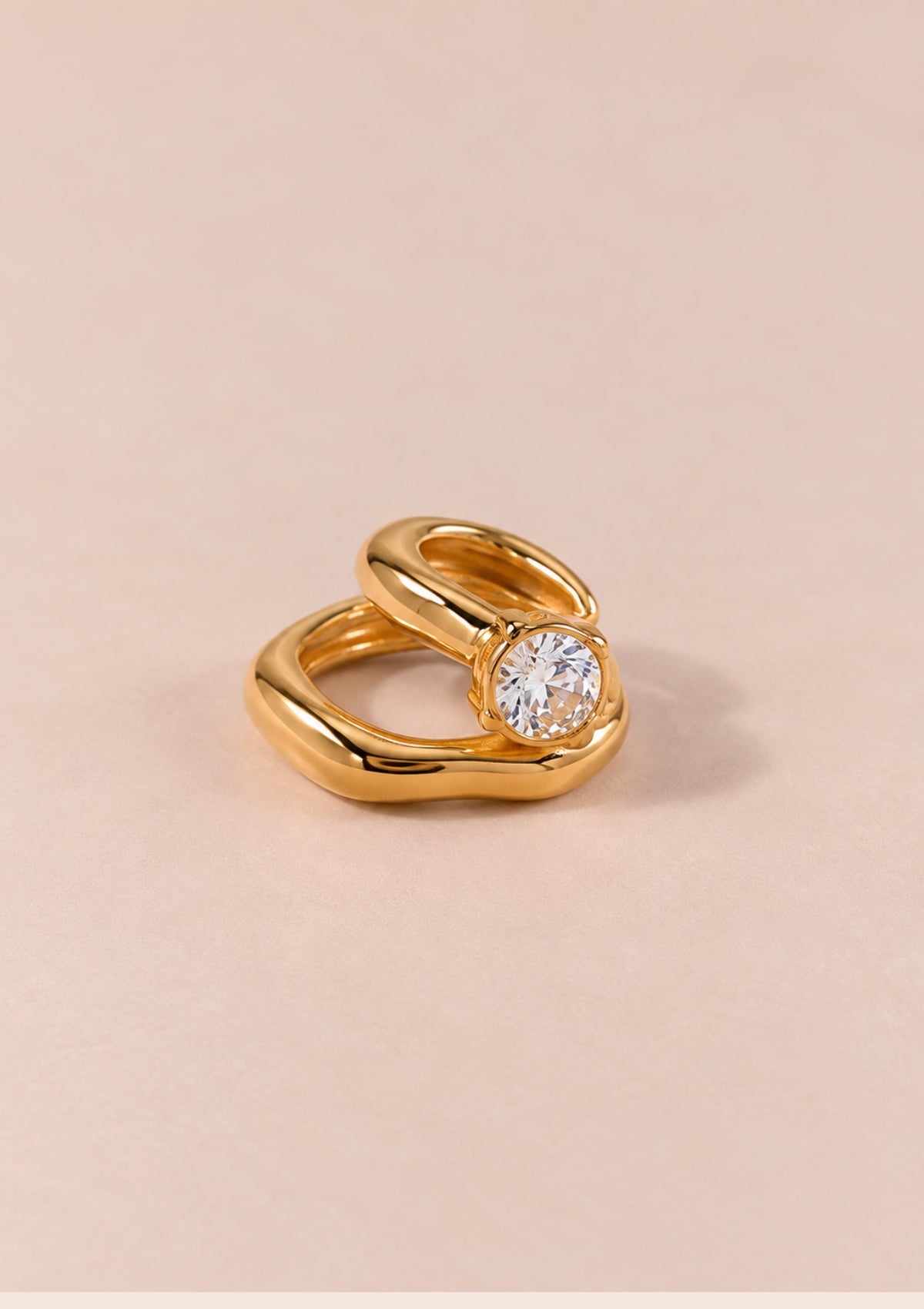 A gold ring with a twisted, fluid design and a round clear gemstone, photographed on a solid pale pink background.