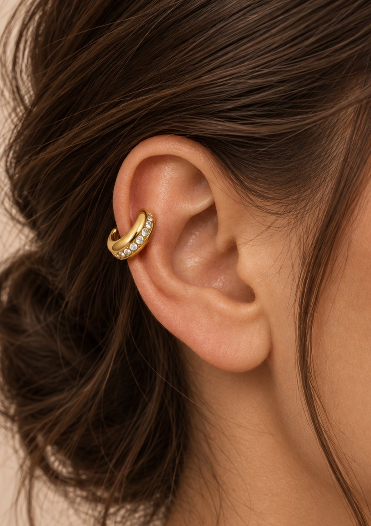 A close-up of a gold huggie hoop earring with small clear stones, worn on a person's ear.