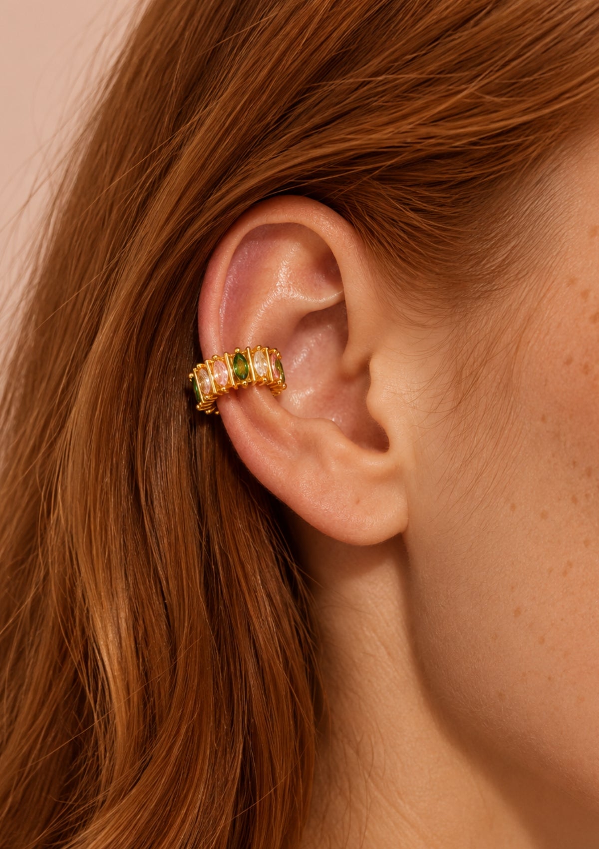 A close-up of a model's ear wearing a gold ear cuff adorned with small clear crystals, set against a soft beige background.