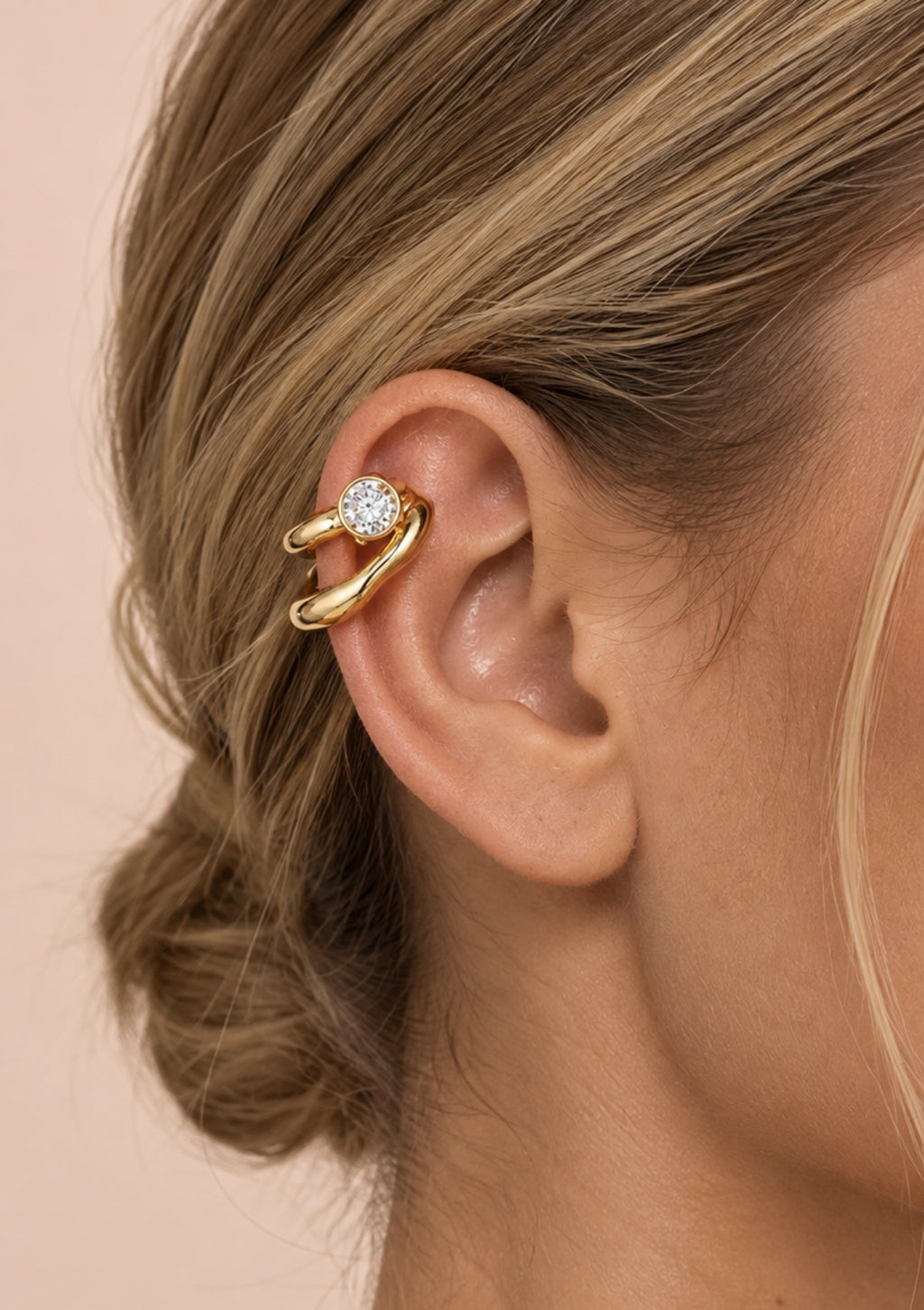 A close-up of a woman's ear wearing a gold ear cuff with a clear round gemstone, set against a soft beige background.