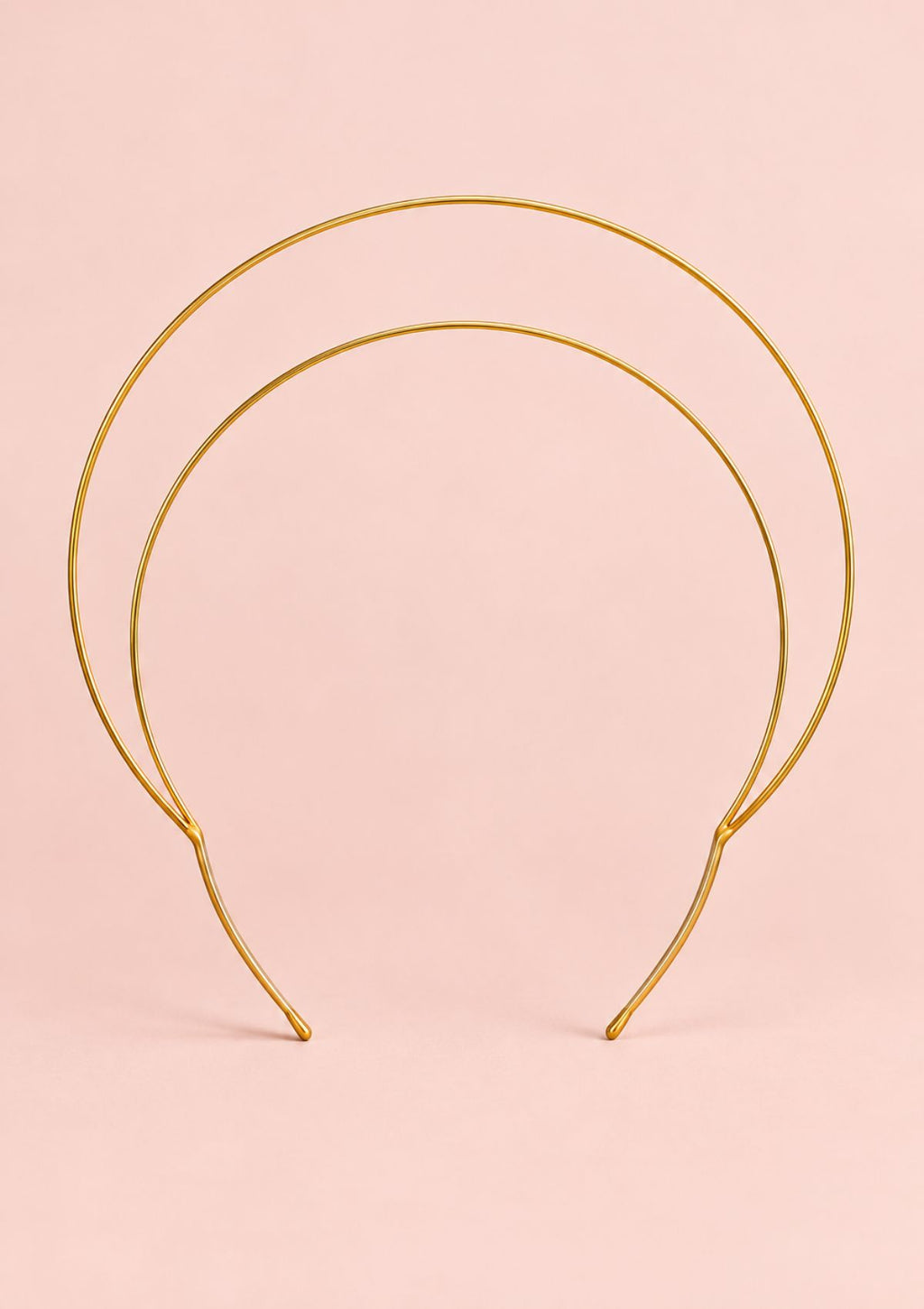 A double-tiered gold wire headband displayed against a solid pale pink background.