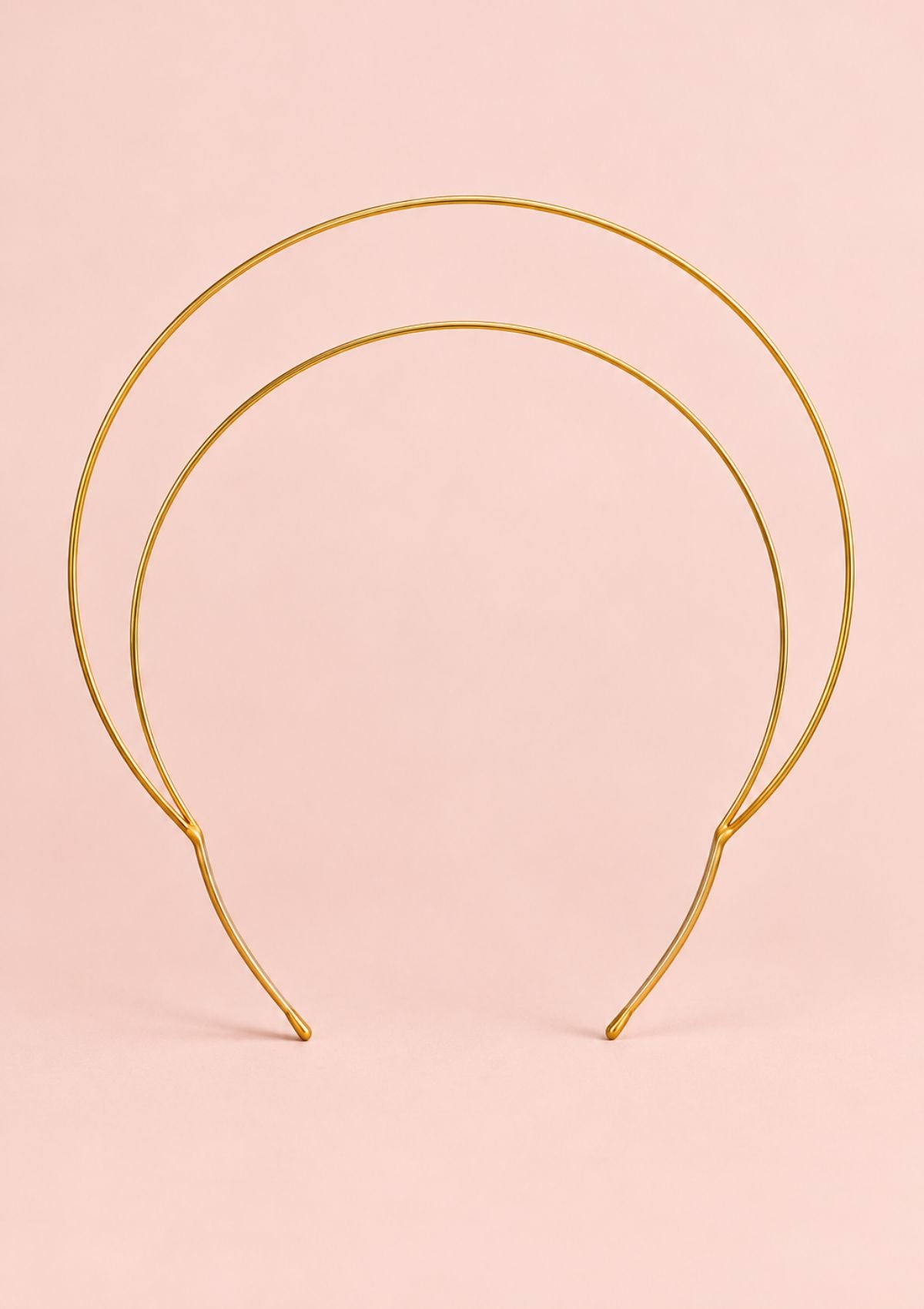 A double-tiered gold wire headband displayed against a solid pale pink background.