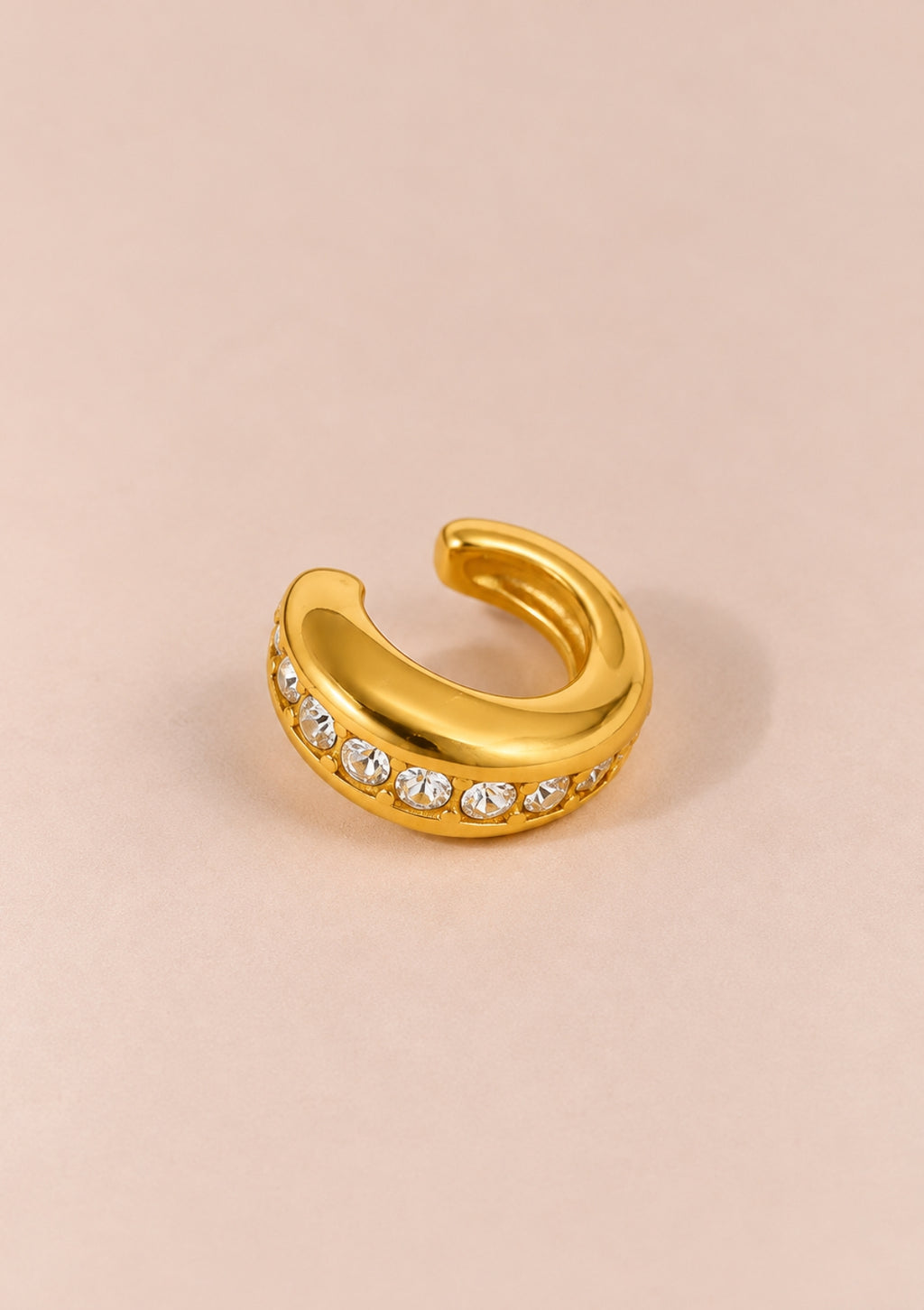 A gold-toned curved ring with a row of clear rhinestones along the front, photographed on a soft beige background.