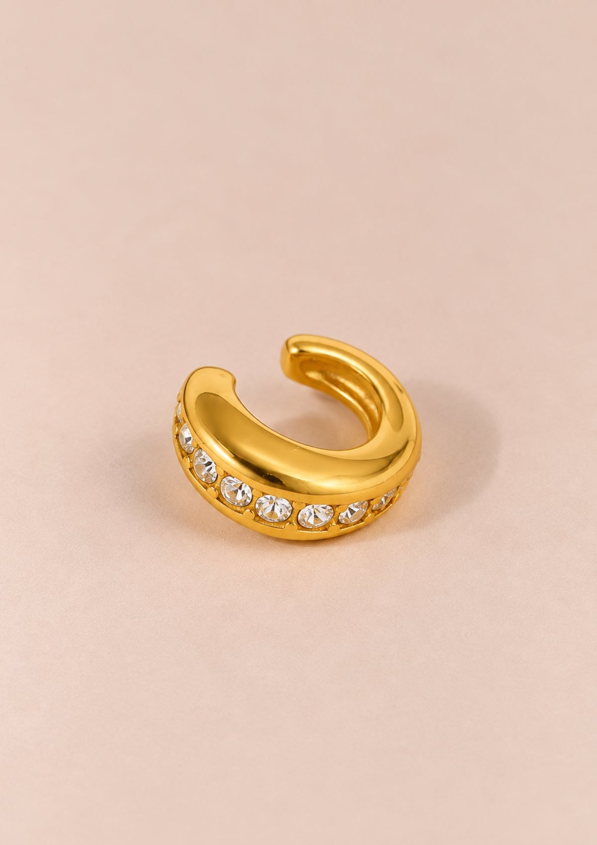 A gold-toned curved ring with a row of clear rhinestones along the front, photographed on a soft beige background.