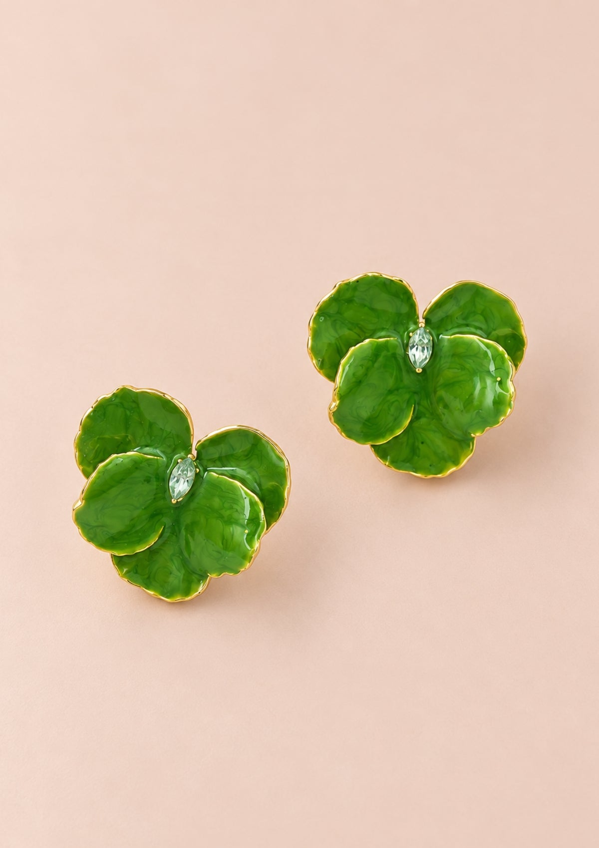 A pair of green enamel four-leaf clover stud earrings with gold trim and a small clear crystal in the center, displayed on a solid pale pink background.