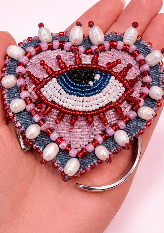 Heart-shaped bracelet with eye design held in a hand against a light pink background