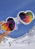 Heart-shaped sunglasses with colorful lenses held up against a snowy mountain background