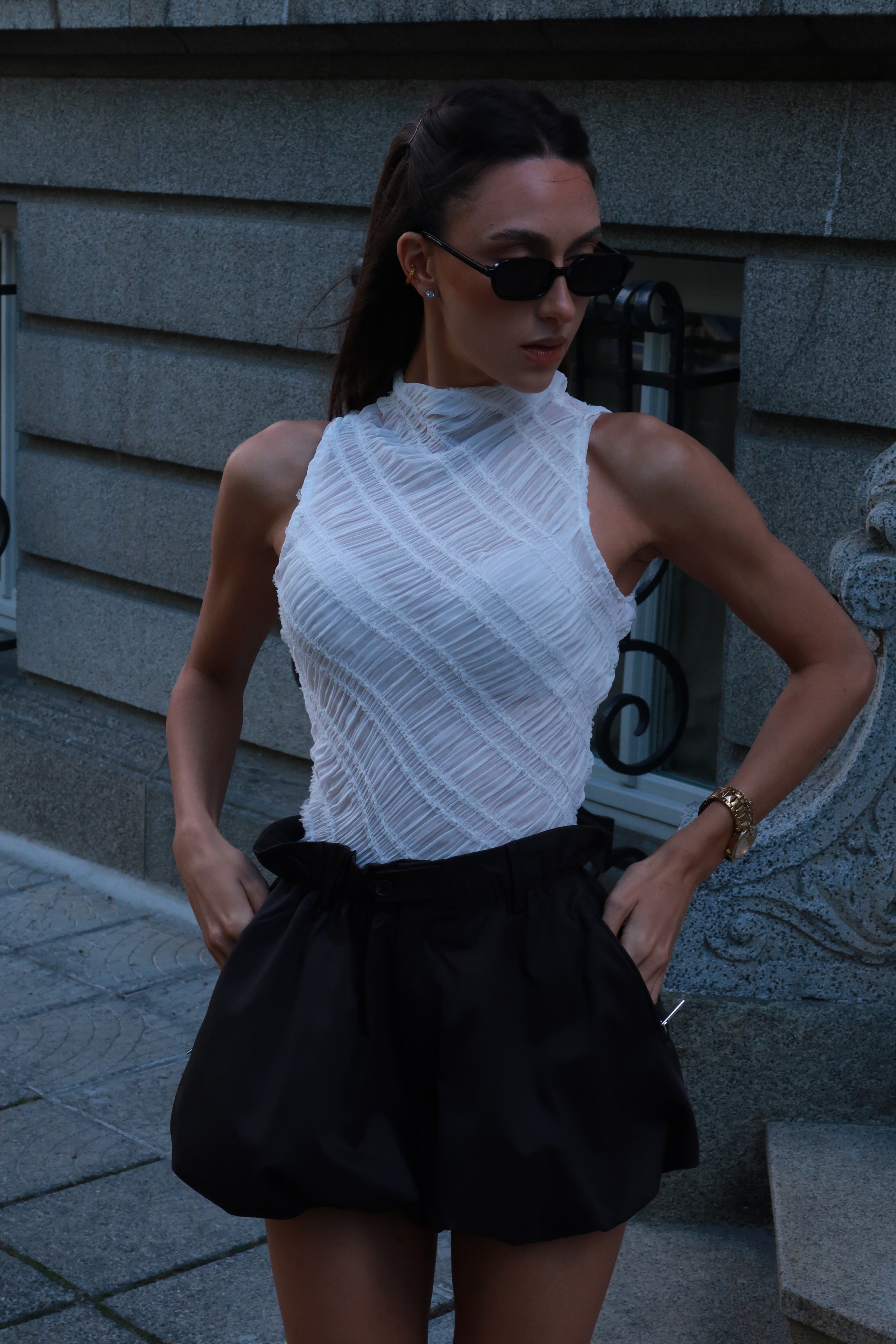 Woman wearing a white sleeveless top and black shorts standing against a gray wall.
