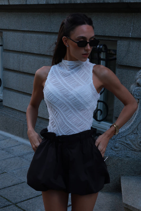 Woman wearing a white sleeveless top and black shorts standing against a gray wall.