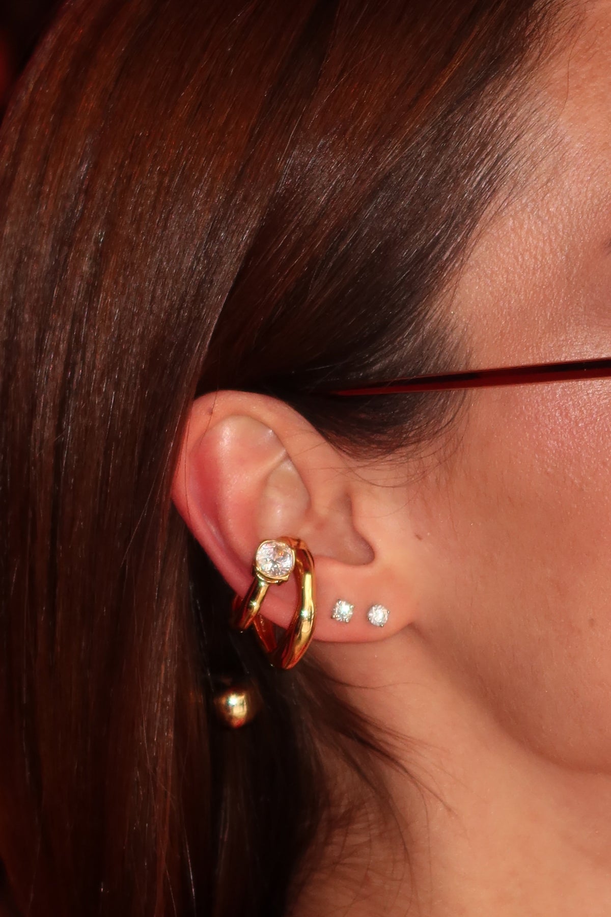A close-up side profile of a woman with dark brown hair wearing a gold hoop earring with a clear gemstone and pearl studs, paired with dark sunglasses and a red top.