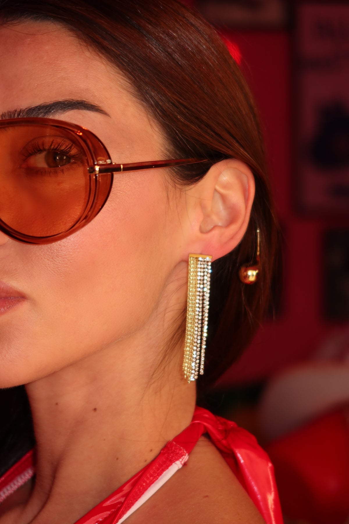 A close-up side profile of a woman wearing a long, dangling rhinestone-encrusted gold earring and a smaller matching hoop earring, paired with amber-tinted sunglasses and a red top.