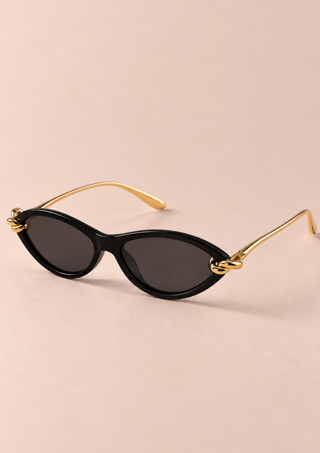 Oval sunglasses with a glossy black frame, dark lenses, and gold metal temples, displayed on a solid pale pink background.