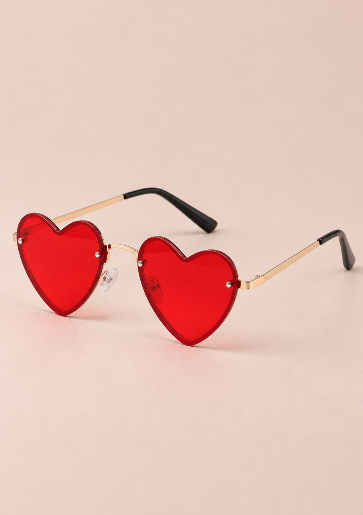 Heart-shaped sunglasses with translucent red lenses and thin gold metal frames, shown on a soft beige background.