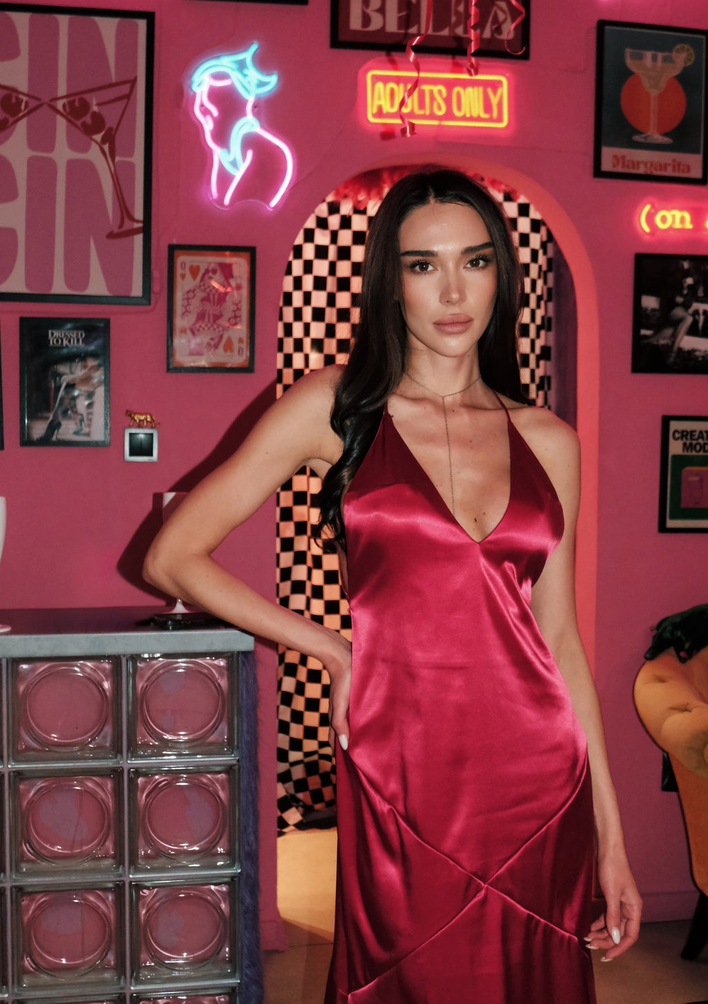Woman in a red dress standing in a room with neon signs and posters on a pink wall.