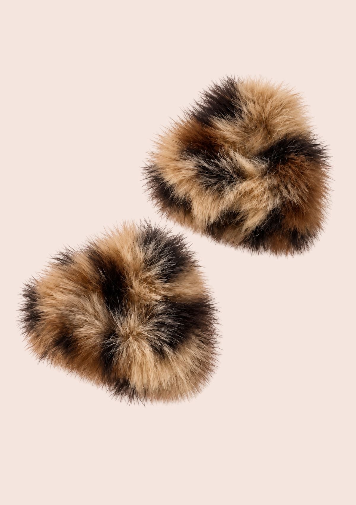 Two round faux fur slippers with a mottled brown, black, and beige pattern, displayed on a solid pale pink background.