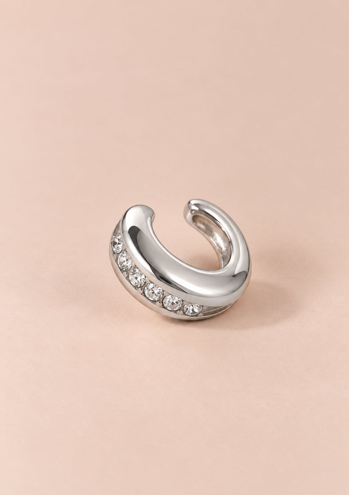 A polished silver-tone crescent-shaped hoop earring with a row of clear gemstones along the front curve, displayed on a solid pale peach-pink background.