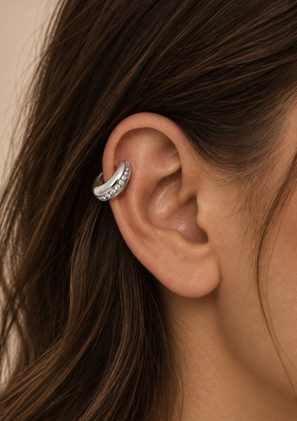 A close-up of a woman's ear wearing a curved silver ear cuff adorned with small clear stones, set against a soft beige background.
