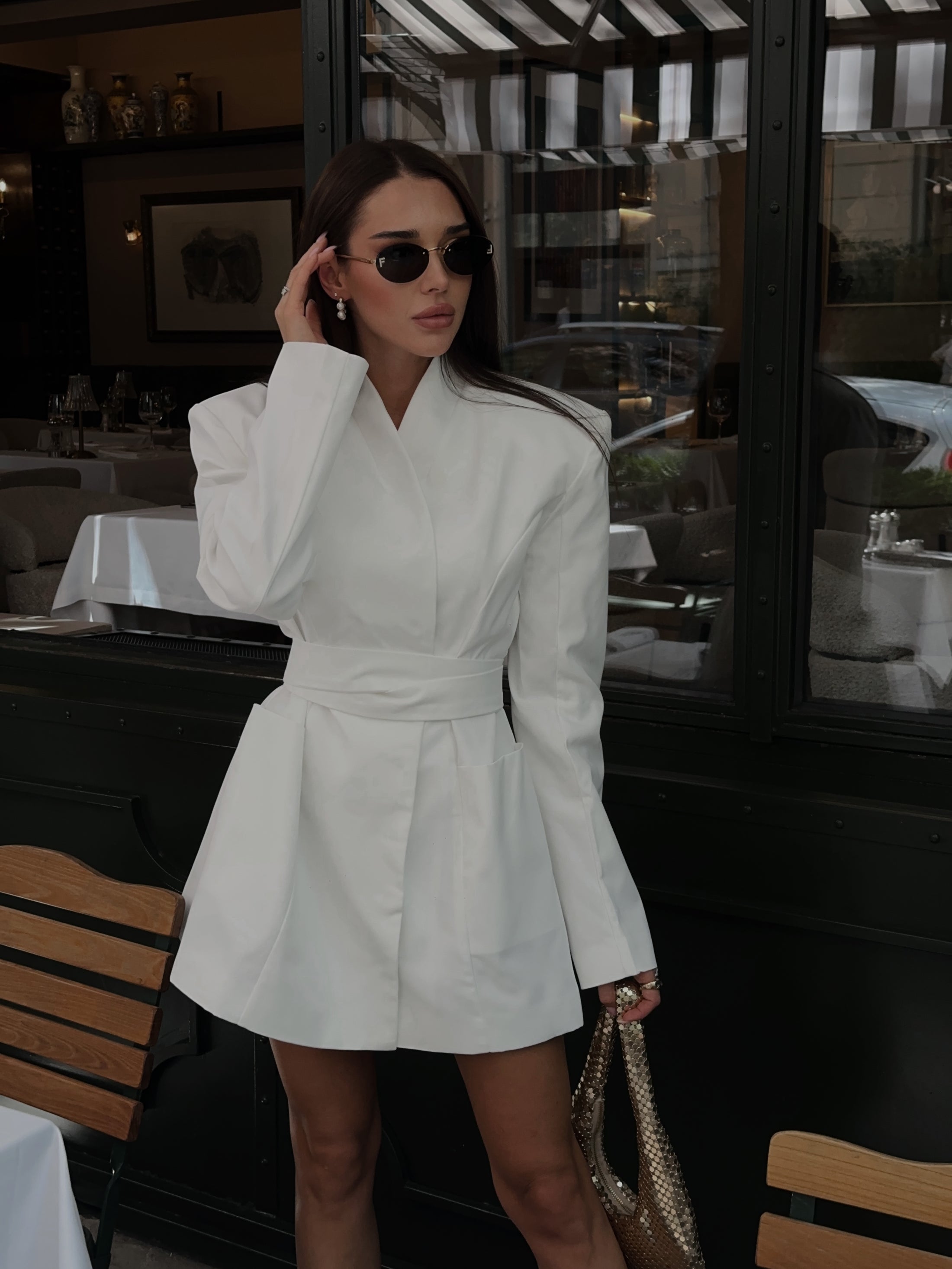 Woman in a white blazer dress with belt standing in an indoor setting with tables and chairs.