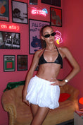 Load image into Gallery viewer, Woman in a black bikini top and white skirt posing in a room with neon signs and framed pictures.
